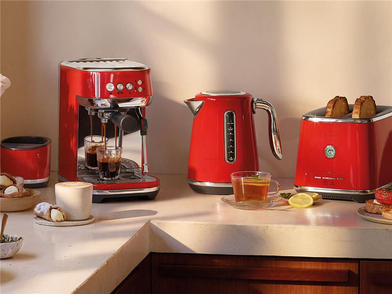 Breville kitchen appliances on a countertop, including an espresso machine pouring coffee, a kettle, and a toaster with toast, styled with coffee, tea, and pastries.